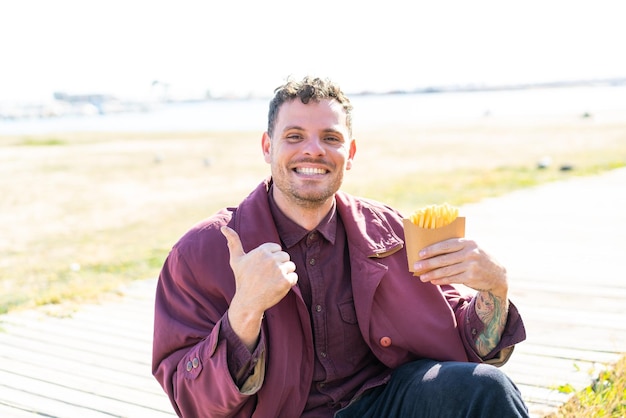 Giovane uomo caucasico che tiene patatine fritte all'aperto indicando il lato per presentare un prodotto