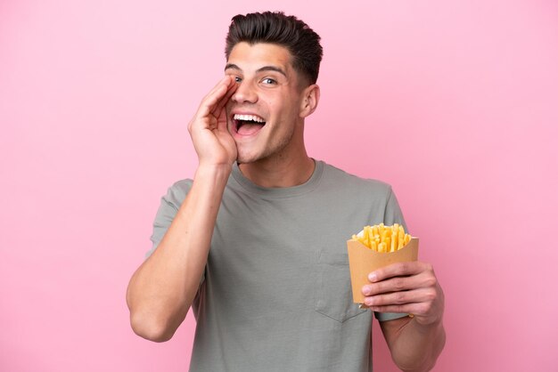 Giovane uomo caucasico che tiene patatine fritte isolate su sfondo rosa che grida con la bocca spalancata