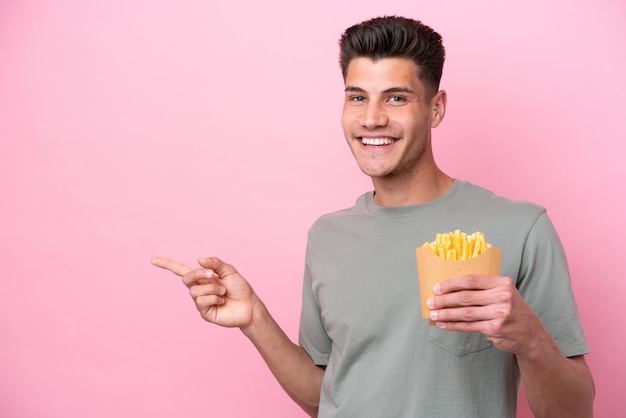 Giovane uomo caucasico che tiene patatine fritte isolate su sfondo rosa che punta il dito di lato