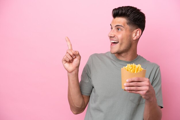 Giovane uomo caucasico che tiene patatine fritte isolate su sfondo rosa con l'intenzione di realizzare la soluzione sollevando un dito