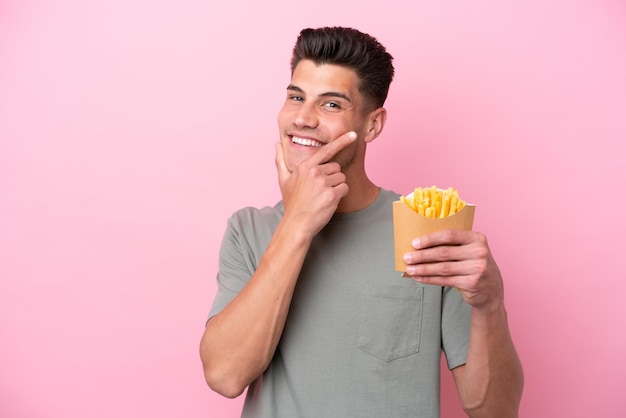 Giovane uomo caucasico che tiene patatine fritte isolate su sfondo rosa felice e sorridente