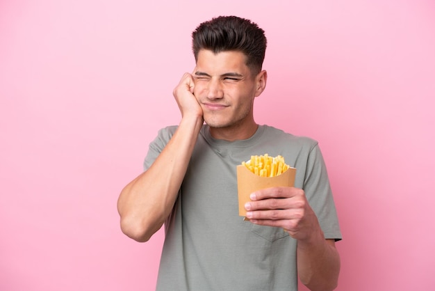 Giovane uomo caucasico che tiene patatine fritte isolate su sfondo rosa frustrato e copre le orecchie