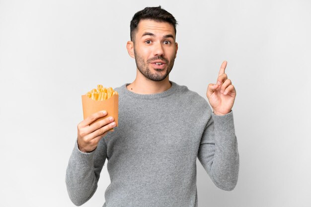 Giovane uomo caucasico che tiene patatine fritte su sfondo bianco isolato pensando un'idea che punta il dito verso l'alto