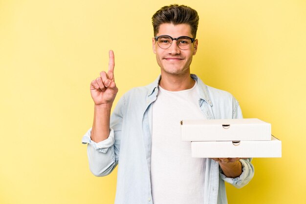 Giovane uomo caucasico che tiene pizze isolate su sfondo giallo che mostra il numero uno con il dito.