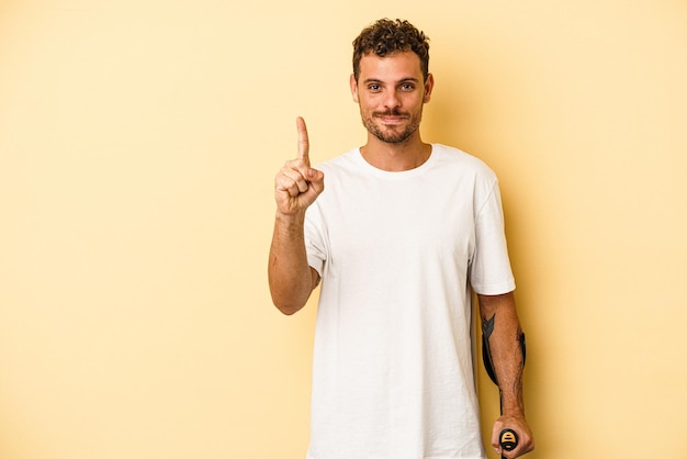 Giovane uomo caucasico che tiene stampella isolata su sfondo giallo che mostra il numero uno con il dito.