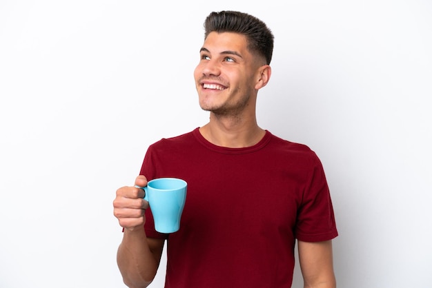 Giovane uomo caucasico che tiene tazza di caffè isolato su sfondo bianco alzando lo sguardo mentre sorride