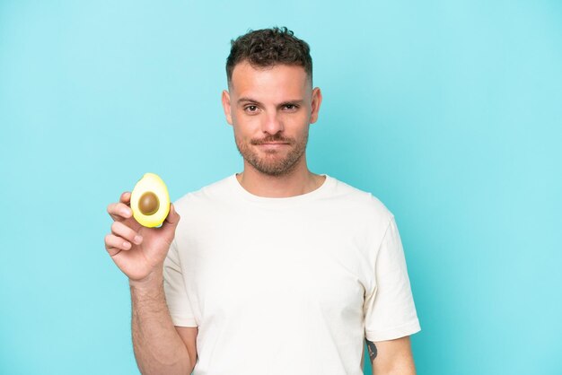 Giovane uomo caucasico che tiene un avocado isolato su sfondo blu con espressione triste