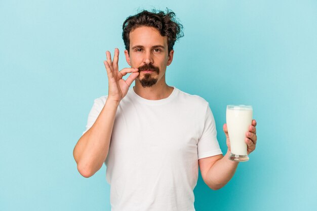 Giovane uomo caucasico che tiene un bicchiere di latte isolato su sfondo blu con le dita sulle labbra mantenendo un segreto.