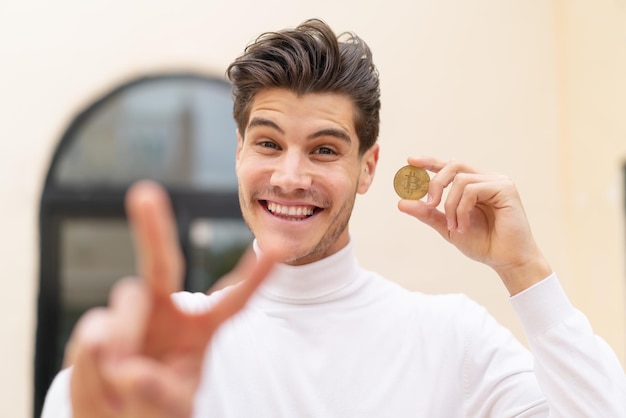 Giovane uomo caucasico che tiene un Bitcoin all'aperto sorridendo e mostrando il segno della vittoria