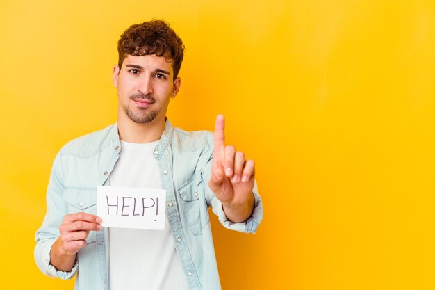 Giovane uomo caucasico che tiene un cartello di aiuto isolato che mostra il numero uno con il dito.