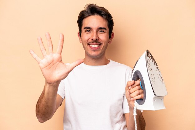Giovane uomo caucasico che tiene un ferro isolato su sfondo beige sorridente allegro che mostra il numero cinque con le dita.