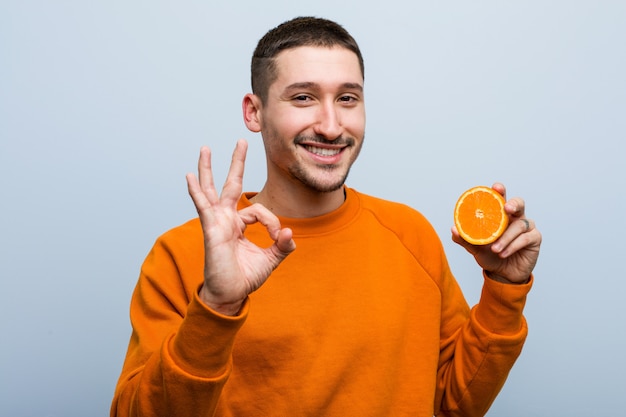 Giovane uomo caucasico che tiene un gesto giusto di mostra allegro e sicuro arancio.