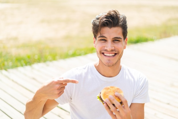 Giovane uomo caucasico che tiene un hamburger all'aperto e lo indica