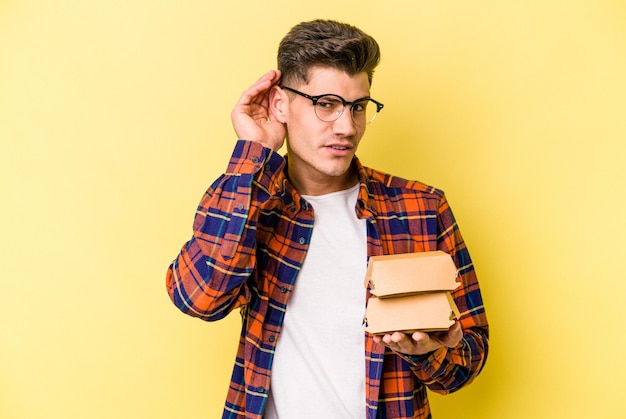 Giovane uomo caucasico che tiene un hamburger isolato su sfondo giallo cercando di ascoltare un pettegolezzo.