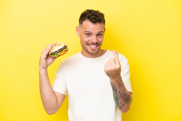 Giovane uomo caucasico che tiene un hamburger isolato su sfondo giallo che fa gesto di denaro