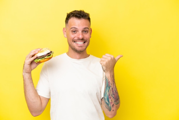 Giovane uomo caucasico che tiene un hamburger isolato su sfondo giallo che indica il lato per presentare un prodotto