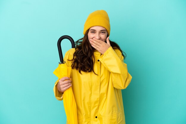 Giovane uomo caucasico che tiene un ombrello isolato su sfondo blu felice e sorridente che copre la bocca con la mano