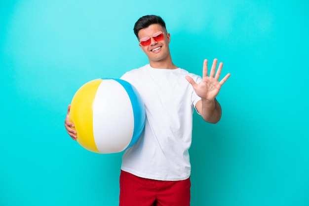 Giovane uomo caucasico che tiene un pallone da spiaggia isolato su sfondo blu contando cinque con le dita