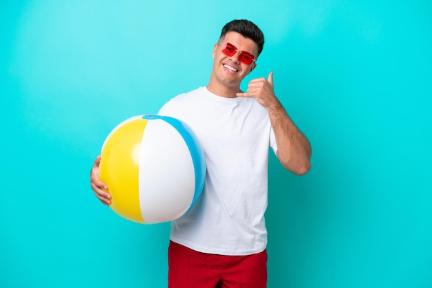 Giovane uomo caucasico che tiene un pallone da spiaggia isolato su sfondo blu facendo il gesto del telefono Chiamami indietro segno