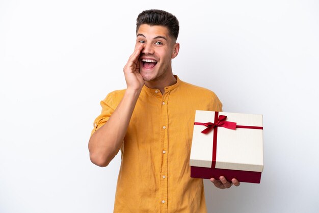 Giovane uomo caucasico che tiene un regalo isolato su sfondo bianco che grida con la bocca spalancata