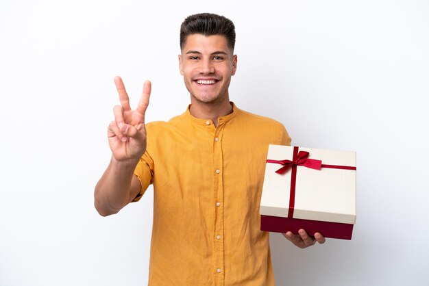 Giovane uomo caucasico che tiene un regalo isolato su sfondo bianco sorridente e mostrando segno di vittoria