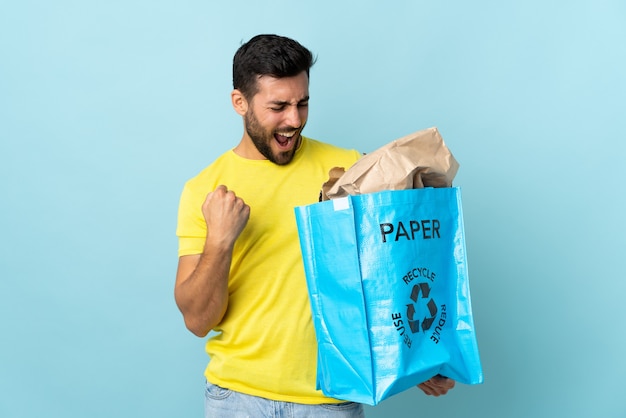 Giovane uomo caucasico che tiene un sacchetto di riciclo isolato su sfondo blu che celebra una vittoria