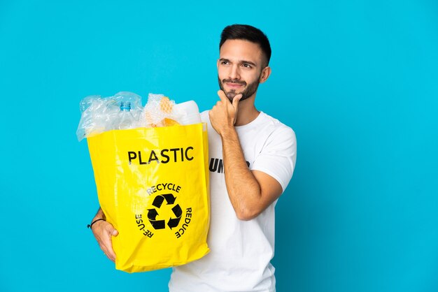 Giovane uomo caucasico che tiene un sacchetto pieno di bottiglie di plastica da riciclare isolato su sfondo blu avendo dubbi e pensando