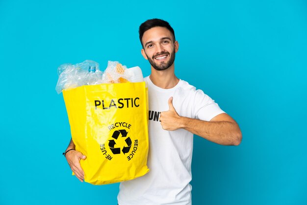 Giovane uomo caucasico che tiene un sacchetto pieno di bottiglie di plastica da riciclare isolato su sfondo blu, dando un pollice in alto gesto