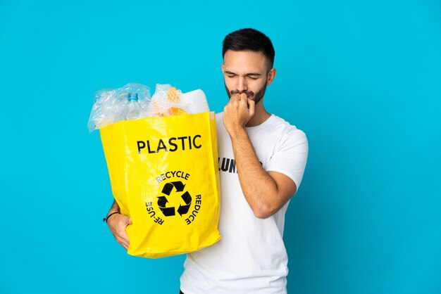 Giovane uomo caucasico che tiene un sacchetto pieno di bottiglie di plastica da riciclare isolato sulla parete blu avendo dubbi
