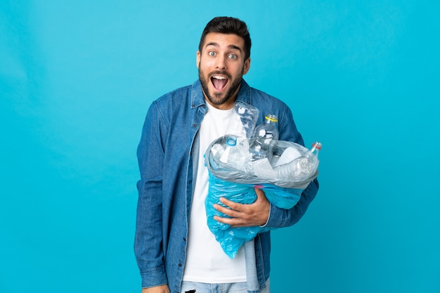 Giovane uomo caucasico che tiene un sacchetto pieno di bottiglie di plastica per riciclare isolato sulla parete blu con sorpresa e scioccata espressione facciale