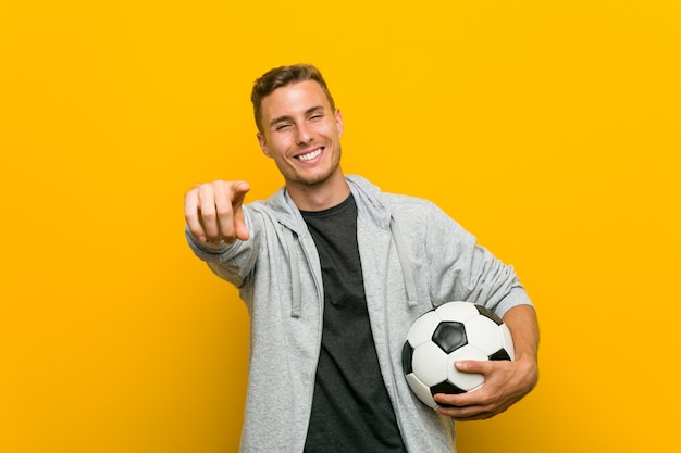 Giovane uomo caucasico che tiene un sorriso allegro di un pallone da calcio che indica la parte anteriore.