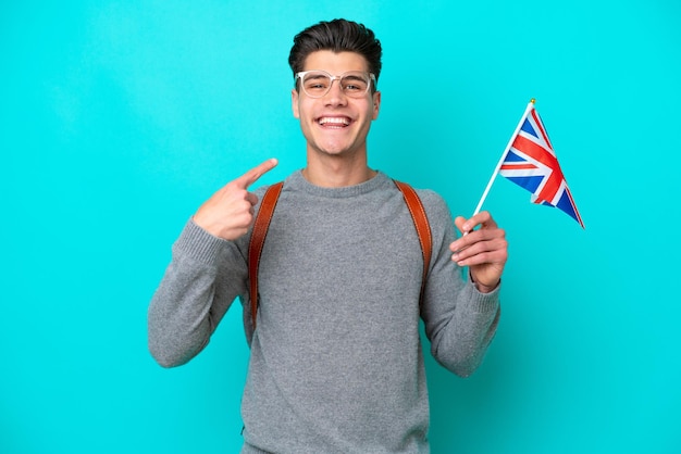 Giovane uomo caucasico che tiene una bandiera del Regno Unito isolata su sfondo blu dando un gesto di pollice in alto