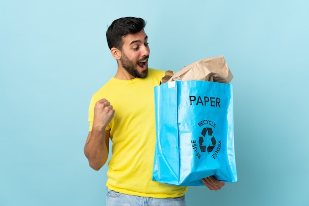 Giovane uomo caucasico che tiene una borsa di riciclaggio isolata sulla parete blu che celebra una vittoria