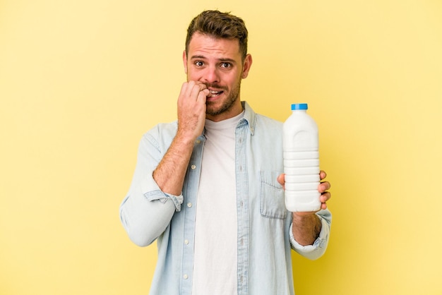 Giovane uomo caucasico che tiene una bottiglia di latte isolata su sfondo giallo che si morde le unghie nervoso e molto ansioso