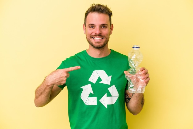 Giovane uomo caucasico che tiene una bottiglia di plastica da riciclare isolata su sfondo giallo persona che indica a mano uno spazio per la copia della maglietta orgoglioso e fiducioso