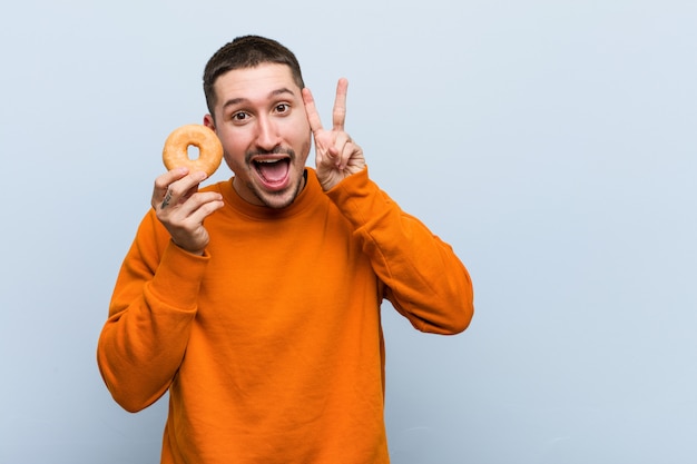 Giovane uomo caucasico che tiene una ciambella che mostra il segno di vittoria e che sorride ampiamente