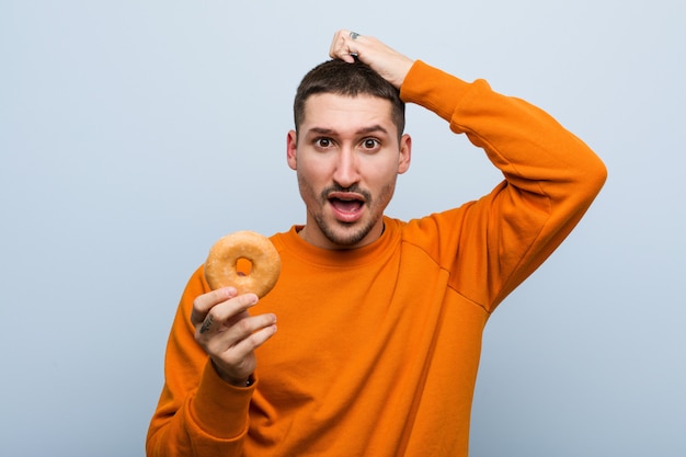 Giovane uomo caucasico che tiene una ciambella scioccata, ha ricordato un incontro importante.