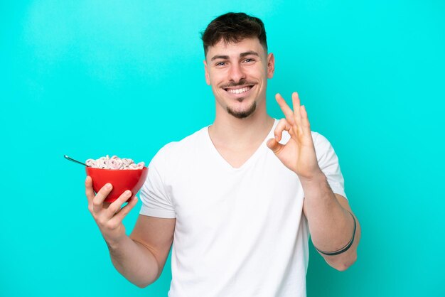 Giovane uomo caucasico che tiene una ciotola di cereali isolata su sfondo blu che mostra il segno giusto con le dita