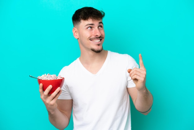 Giovane uomo caucasico che tiene una ciotola di cereali isolata su sfondo blu con l'intenzione di realizzare la soluzione sollevando un dito
