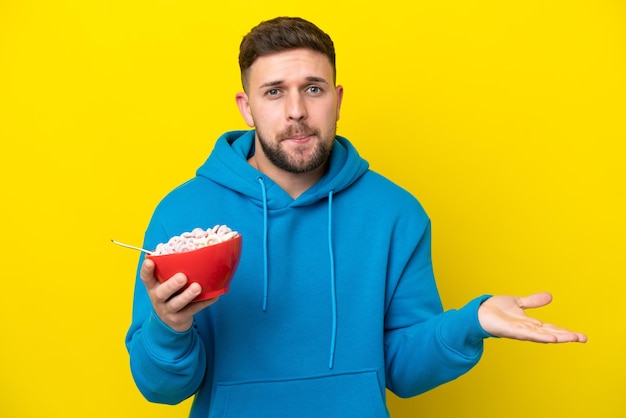 Giovane uomo caucasico che tiene una ciotola di cereali isolata su sfondo giallo facendo un gesto di dubbi mentre si sollevano le spalle