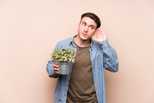 Giovane uomo caucasico che tiene una pianta che prova ad ascoltare un pettegolezzo.