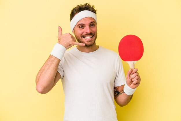 Giovane uomo caucasico che tiene una racchetta da ping pong isolata su sfondo giallo che mostra un gesto di telefonata con le dita.