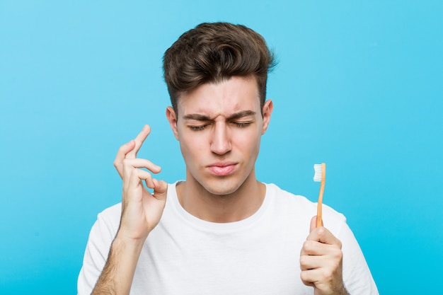 Giovane uomo caucasico che tiene uno spazzolino da denti Giovane donna indiana che indossa un pigiama e una maschera per il sonno isolata tenendo un cuscino incrociando le dita per avere fortuna <mixto>