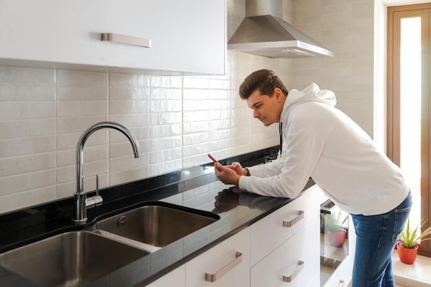 Giovane uomo caucasico che usa il cellulare in cucina È appoggiato al piano di lavoro Indossa una felpa bianca e jeans Cucina luminosa con marmo nero Sta entrando luce naturale