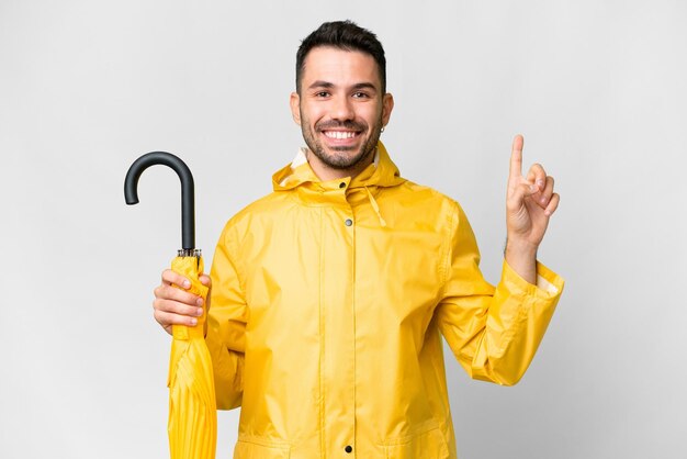 Giovane uomo caucasico con cappotto antipioggia e ombrello isolato su sfondo bianco che mostra e alza un dito in segno del meglio