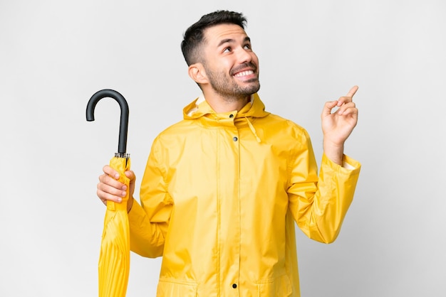 Giovane uomo caucasico con cappotto antipioggia e ombrello isolato su sfondo bianco rivolto verso l'alto una grande idea