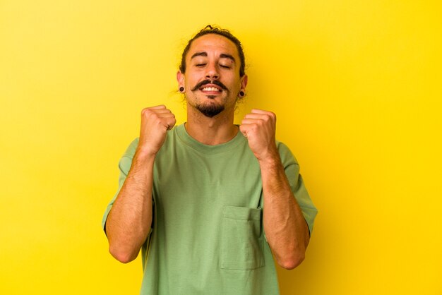 Giovane uomo caucasico con i capelli lunghi isolato su sfondo giallo che celebra una vittoria, passione ed entusiasmo, espressione felice.