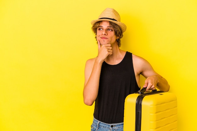 Giovane uomo caucasico con il trucco che va a viaggiare isolato su sfondo giallo guardando di traverso con espressione dubbiosa e scettica.