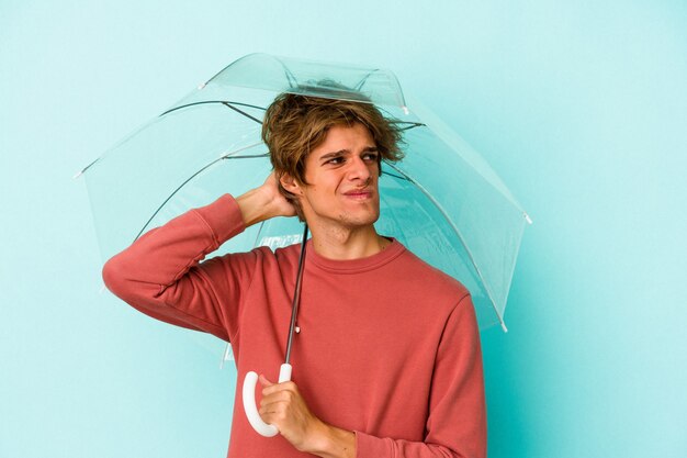 Giovane uomo caucasico con l'ombrello della tenuta di trucco isolato su fondo blu che tocca la parte posteriore della testa, pensando e facendo una scelta.