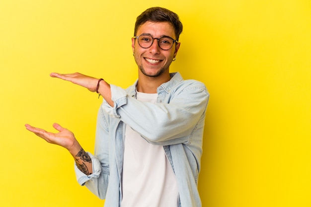 Giovane uomo caucasico con tatuaggi isolati su sfondo giallo che tiene qualcosa di piccolo con l'indice, sorridente e fiducioso.